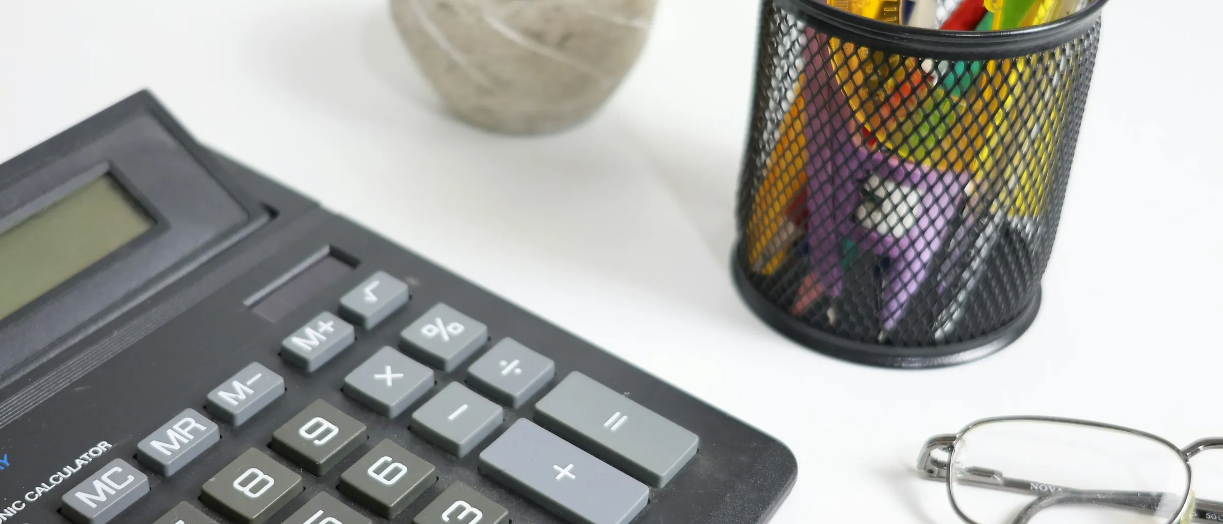 A calculator, reading glasses and pens on a white desk